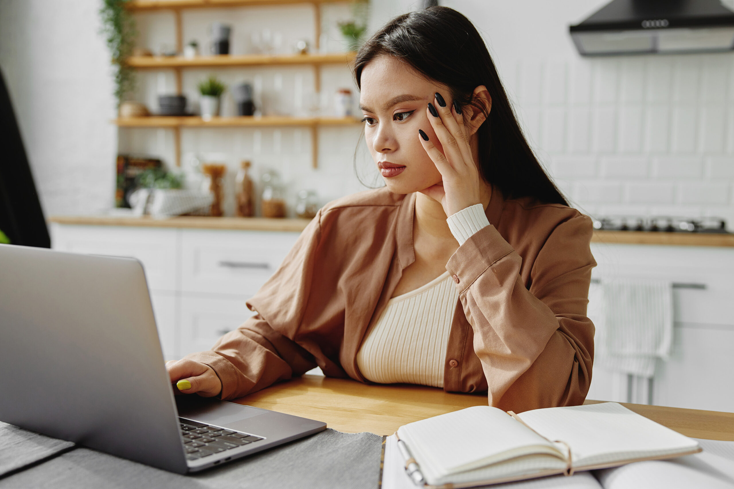 woman sitting at a table, working on her laptop with one hand propped on the side of her face. She has an open notebook beside her laptop.